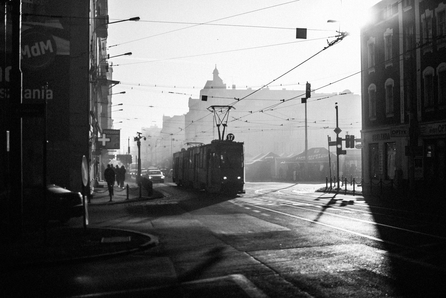 Black and white photo of an urban street scene in Poland. There are buildings, cars, pedestrians, and a tram running on steel rails with an overhead wire for power.